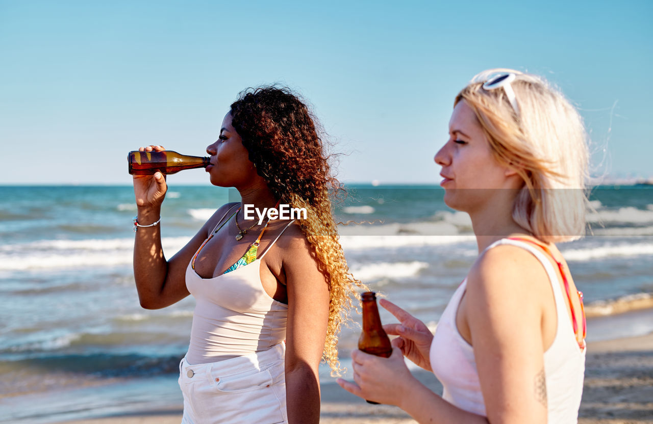 Side view of young multiethnic female friends in summer outfits drinking beer and admiring wavy sea while standing together on sandy beach against cloudless blue sky