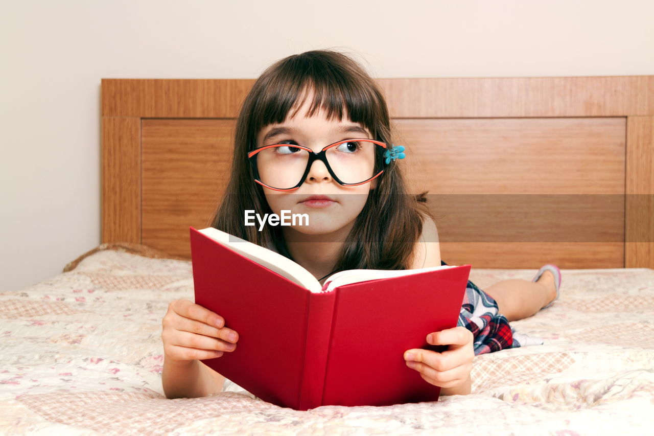 one person, child, childhood, eyeglasses, education, indoors, book, publication, learning, glasses, reading, furniture, portrait, studying, student, women, activity, front view, sitting, intelligence, female, domestic room, bed, relaxation, bedroom, holding, lifestyles, hairstyle, home interior, cute, looking at camera, brown hair, looking, homework, person, domestic life, concentration, emotion, fun, lying down, copy space, innocence, bangs, school, writing, headshot