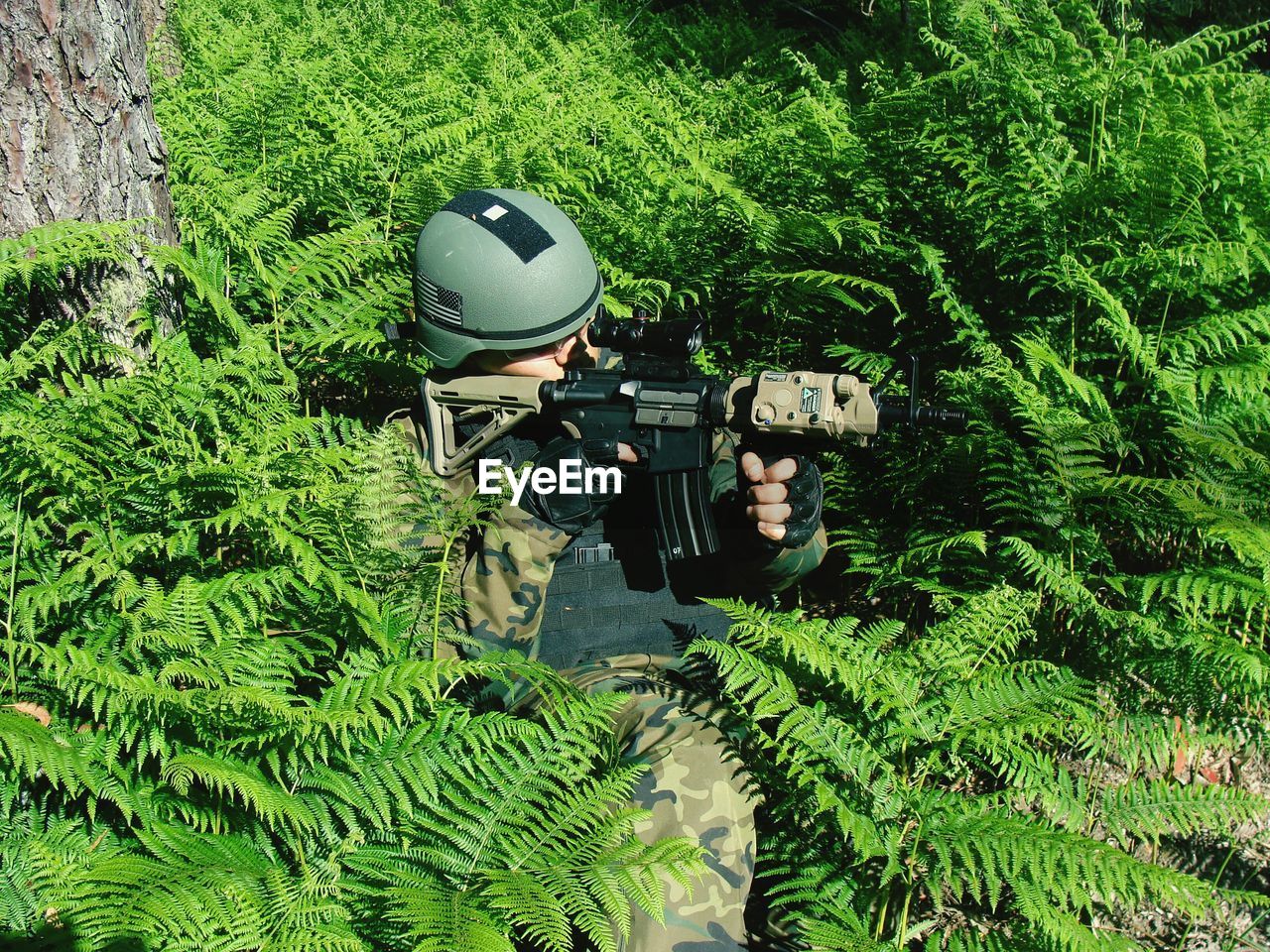 Male soldier aiming rifle while sitting amidst ferns