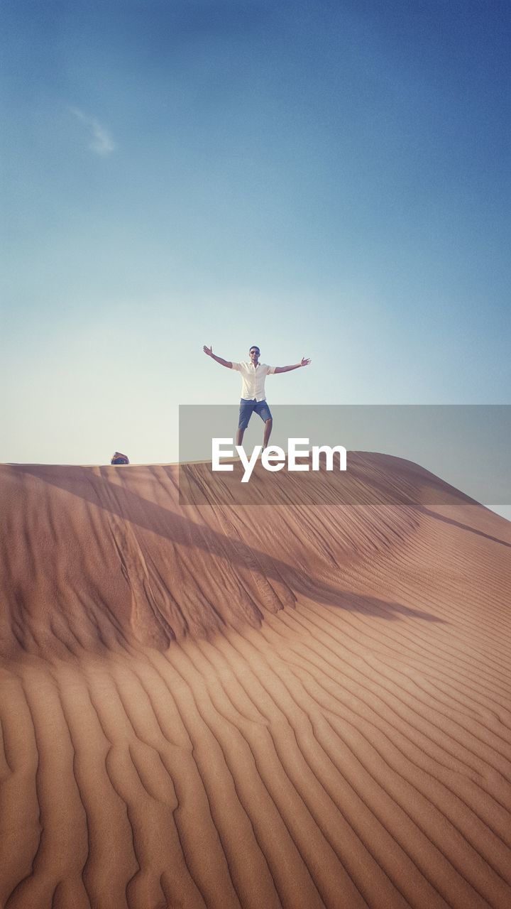 Mid distance view of man with arms outstretched standing on sand dune against blue sky