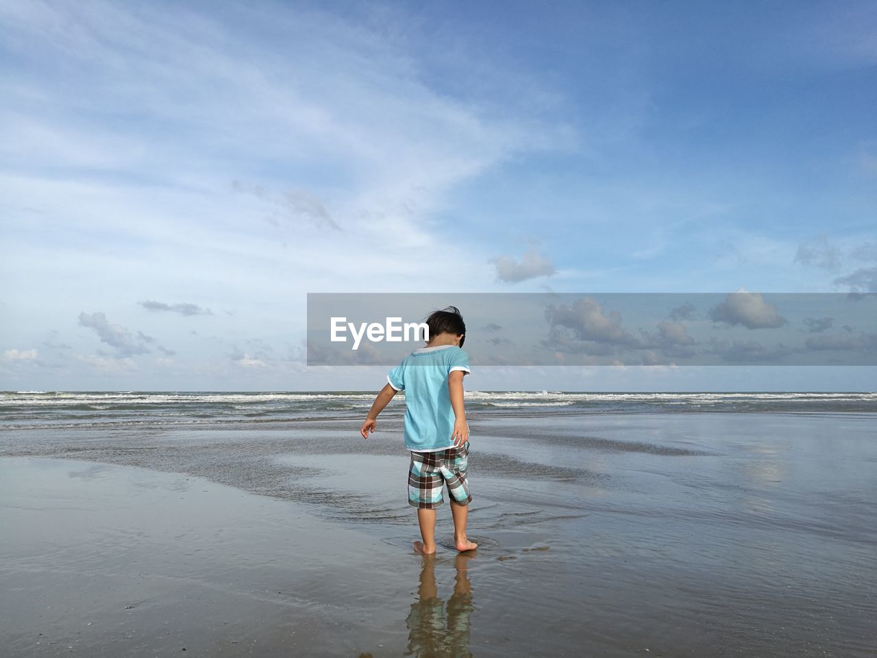 Rear view of boy standing on shore at beach against cloudy sky