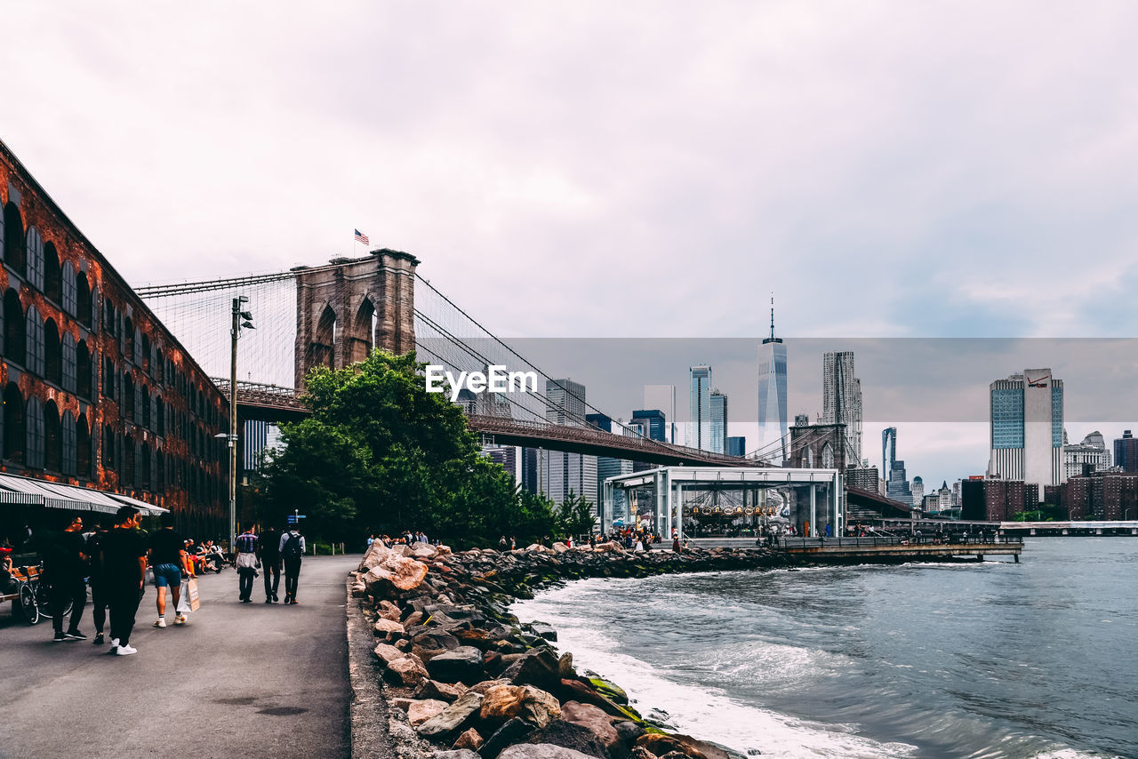 People on street by buildings and brooklyn bridge against cloudy sky