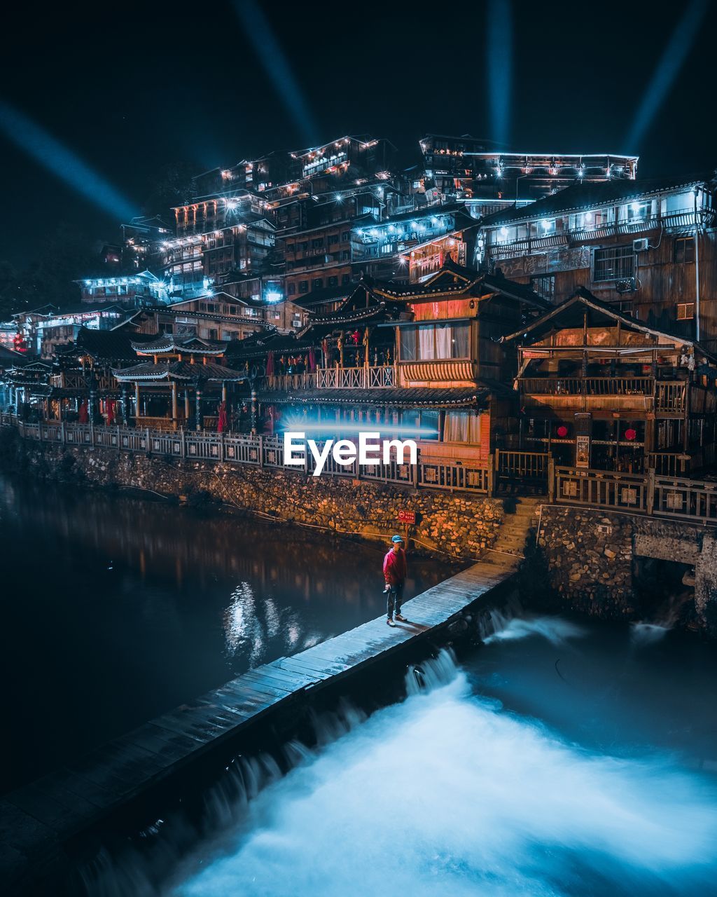 MAN STANDING BY ILLUMINATED RIVER AMIDST BUILDINGS AT NIGHT