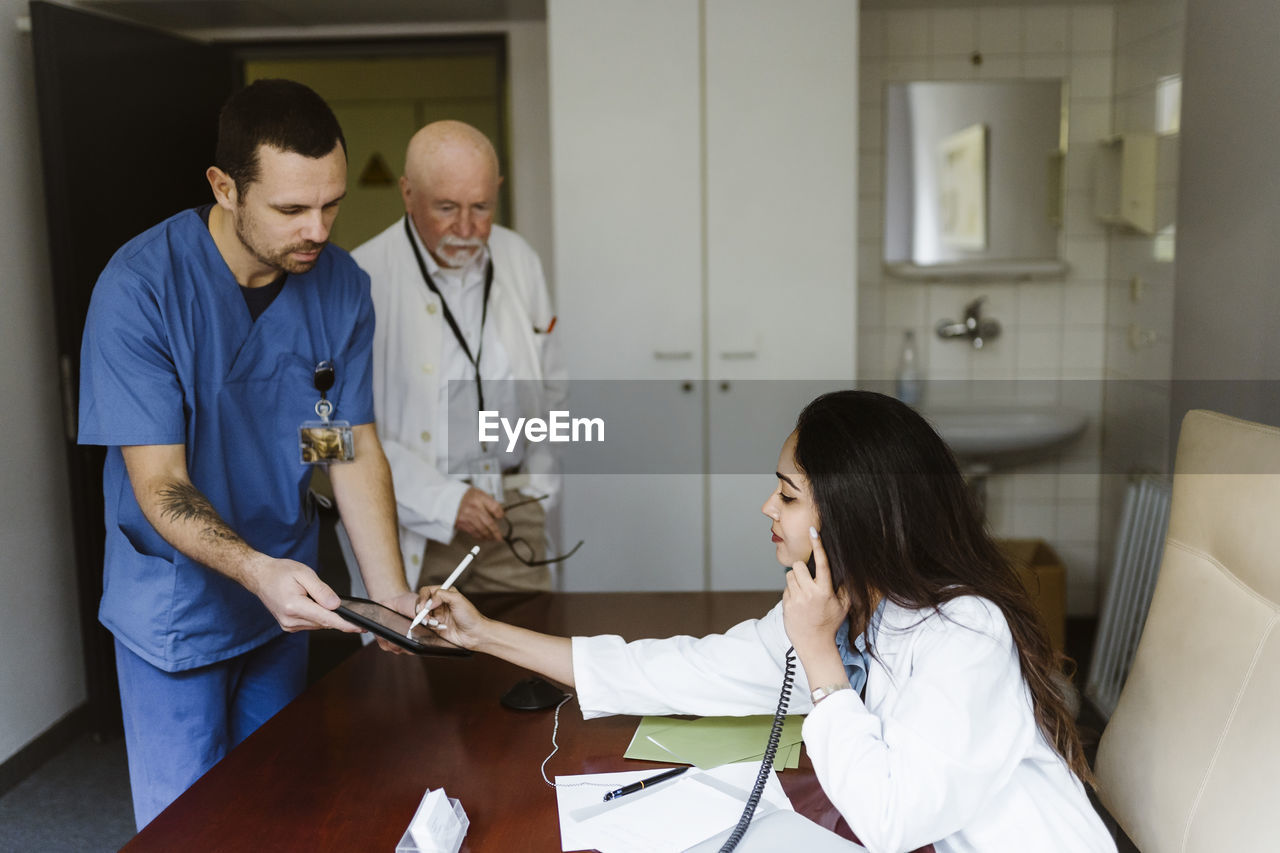 Female doctor talking through telephone while signing on digital tablet held by nurse in office at hospital