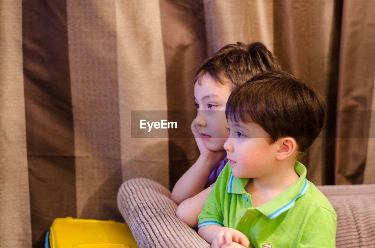 Brothers looking away while sitting on sofa at home