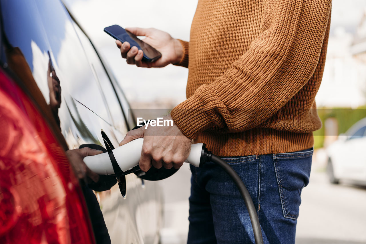 Man with smart phone plugging in electric plug at vehicle charging station