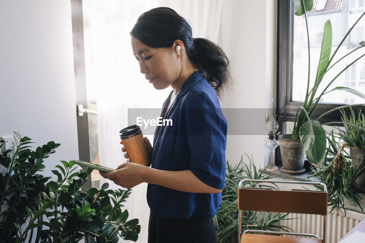 Side view of female architect with reusable coffee cup using smart phone at home office