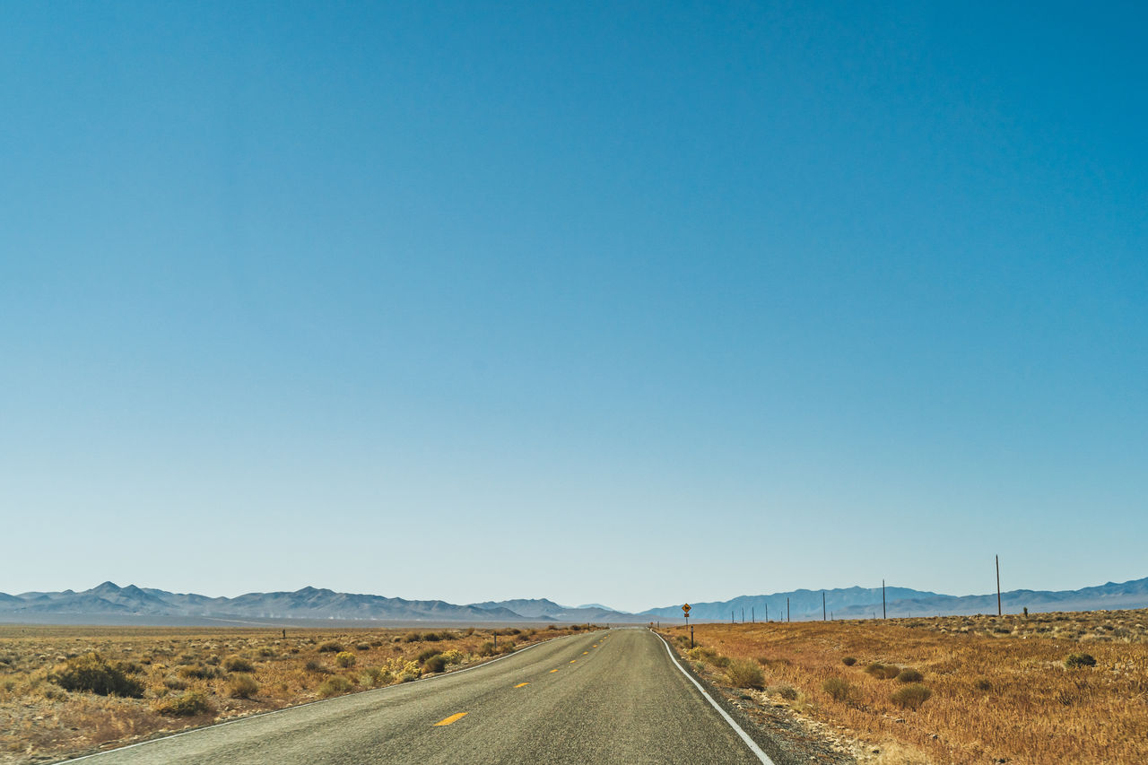 Two lane desert highway road leading to the horizon against clear blue sky and mountains