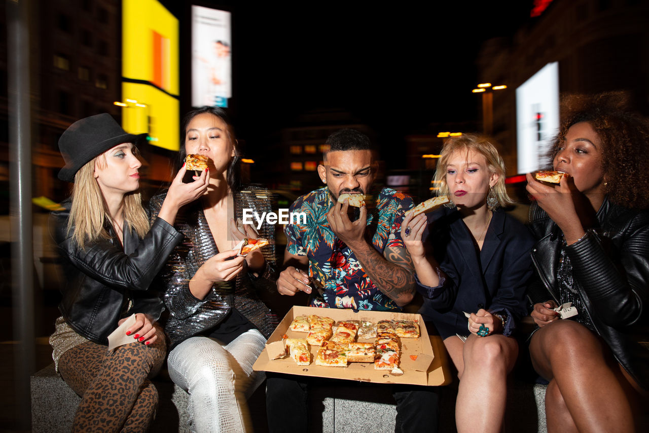 Young multiracial partners with crossed legs enjoying tasty baked food from takeaway box during celebration in town in evening