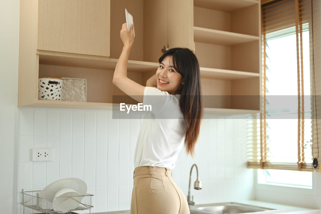one person, women, adult, indoors, domestic room, room, lifestyles, domestic life, home interior, young adult, home, sink, standing, hairstyle, clothing, long hair, bathroom, female, arm, person, emotion, happiness, smiling, interior design, limb, window, waist up, household equipment, human limb, three quarter length, black hair, furniture, arms raised, holding, cheerful, looking, human leg, brown hair, relaxation