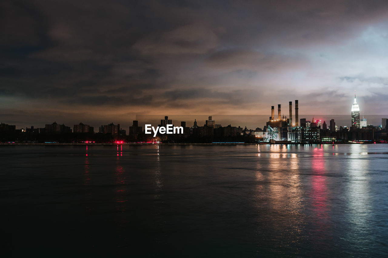 Illuminated buildings by river against sky during sunset