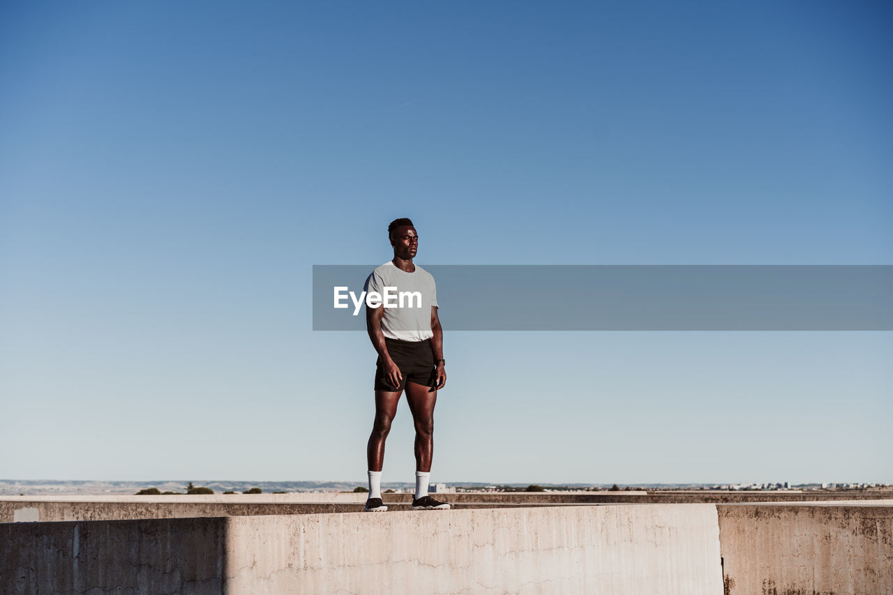 Male athlete standing on wall against blue sky during sunny day