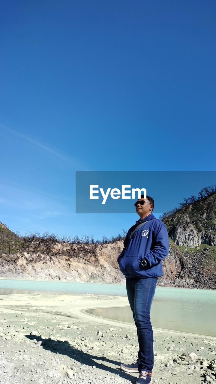 one person, blue, full length, sky, nature, standing, adult, land, sea, vacation, young adult, beach, leisure activity, clear sky, day, copy space, sunny, casual clothing, sunlight, winter, portrait, men, clothing, water, fashion, scenics - nature, lifestyles, beauty in nature, glasses, footwear, environment, sunglasses, outdoors, landscape, shore, holiday, sand, shadow, looking, front view, trip, looking at camera, coast, travel, mountain, non-urban scene