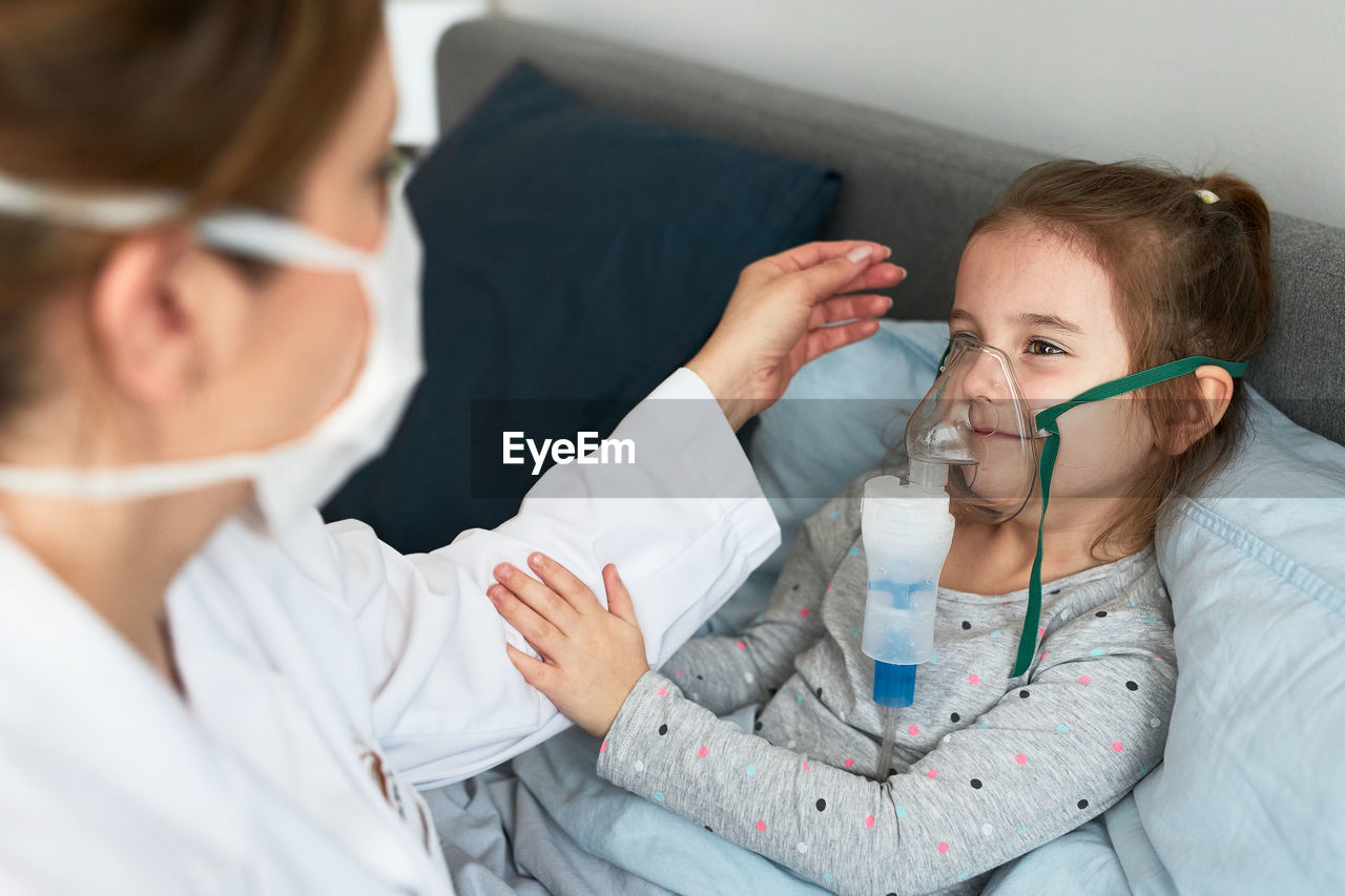 High angle view of female doctor wearing mask examining girl sitting on bed