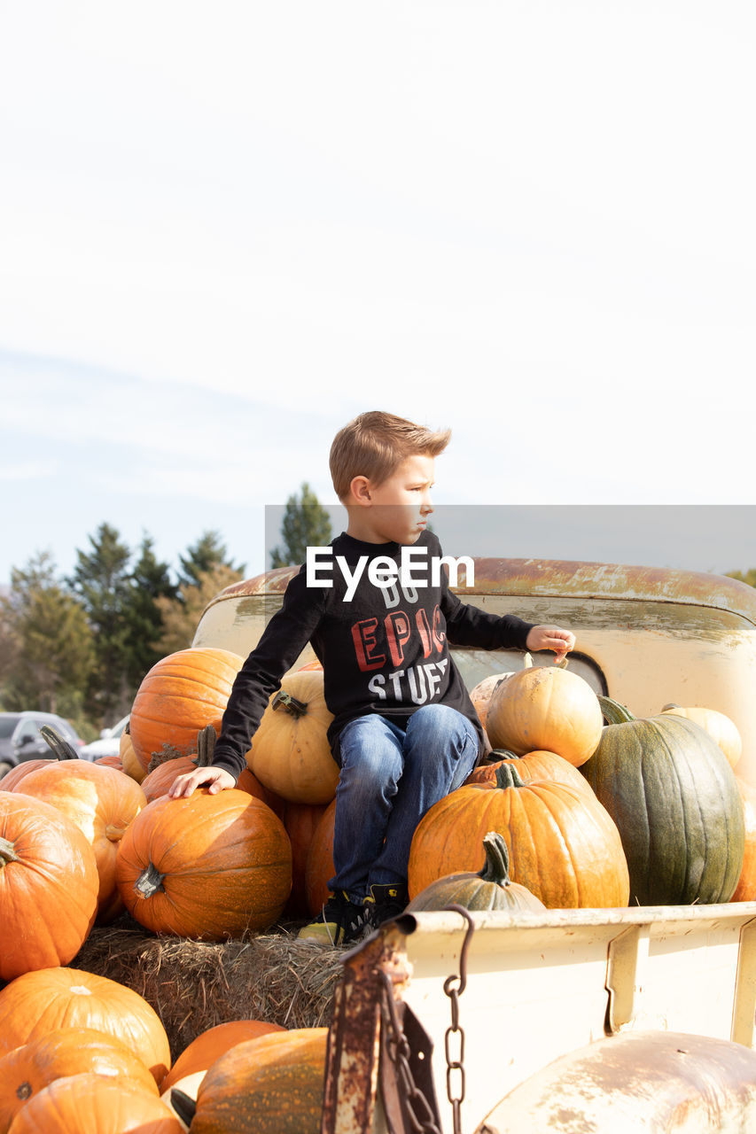 FULL LENGTH OF BOY SITTING ON PUMPKIN AGAINST SKY DURING HALLOWEEN