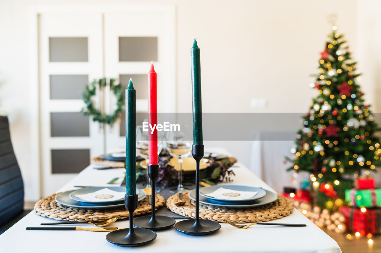Festive table setting with candles located near fancy christmas tree with shiny garlands in spacious living room before family gathering
