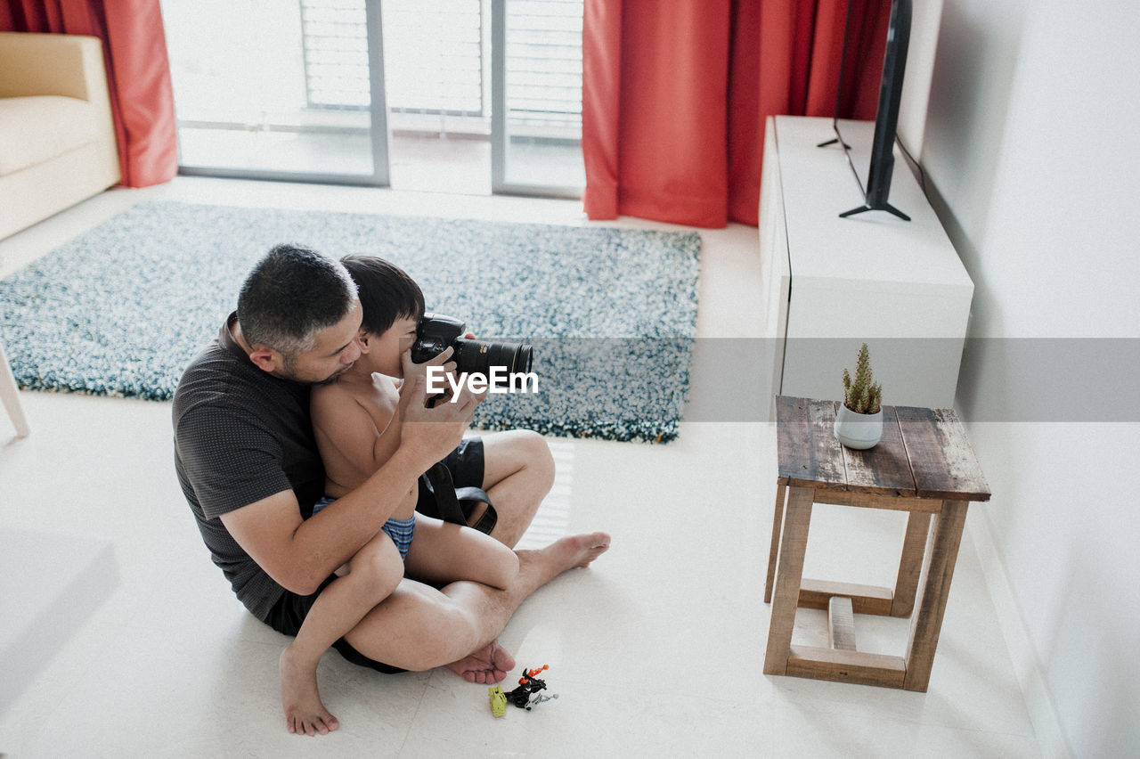 High angle view of father with son photographing while sitting on floor at home