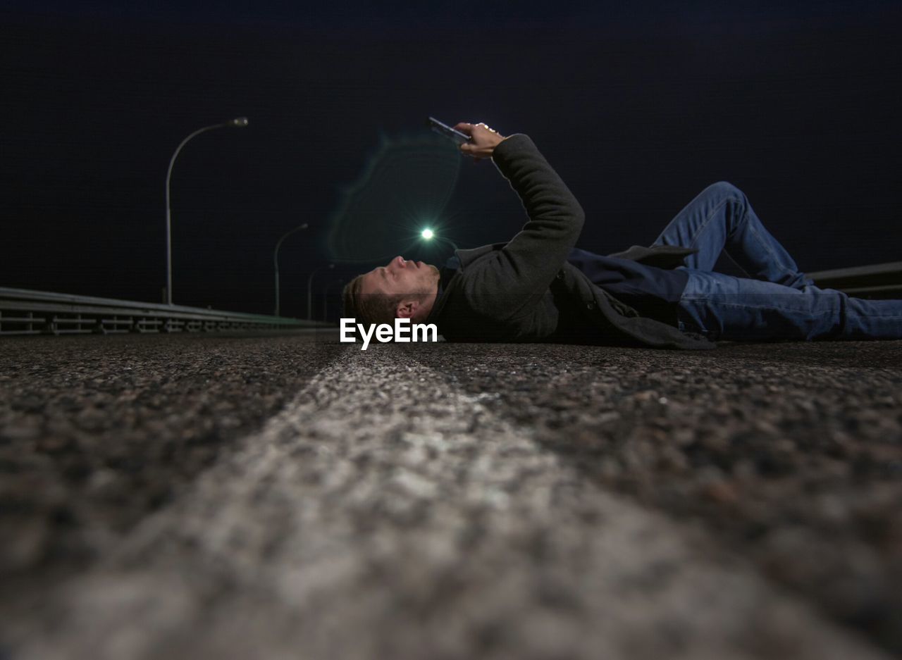 Surface level of young man using mobile phone while lying on street against sky at night