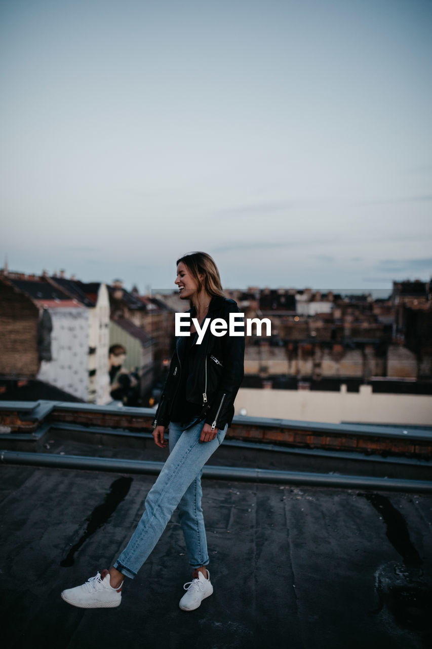 Full length of happy young woman standing on building terrace against sky in city