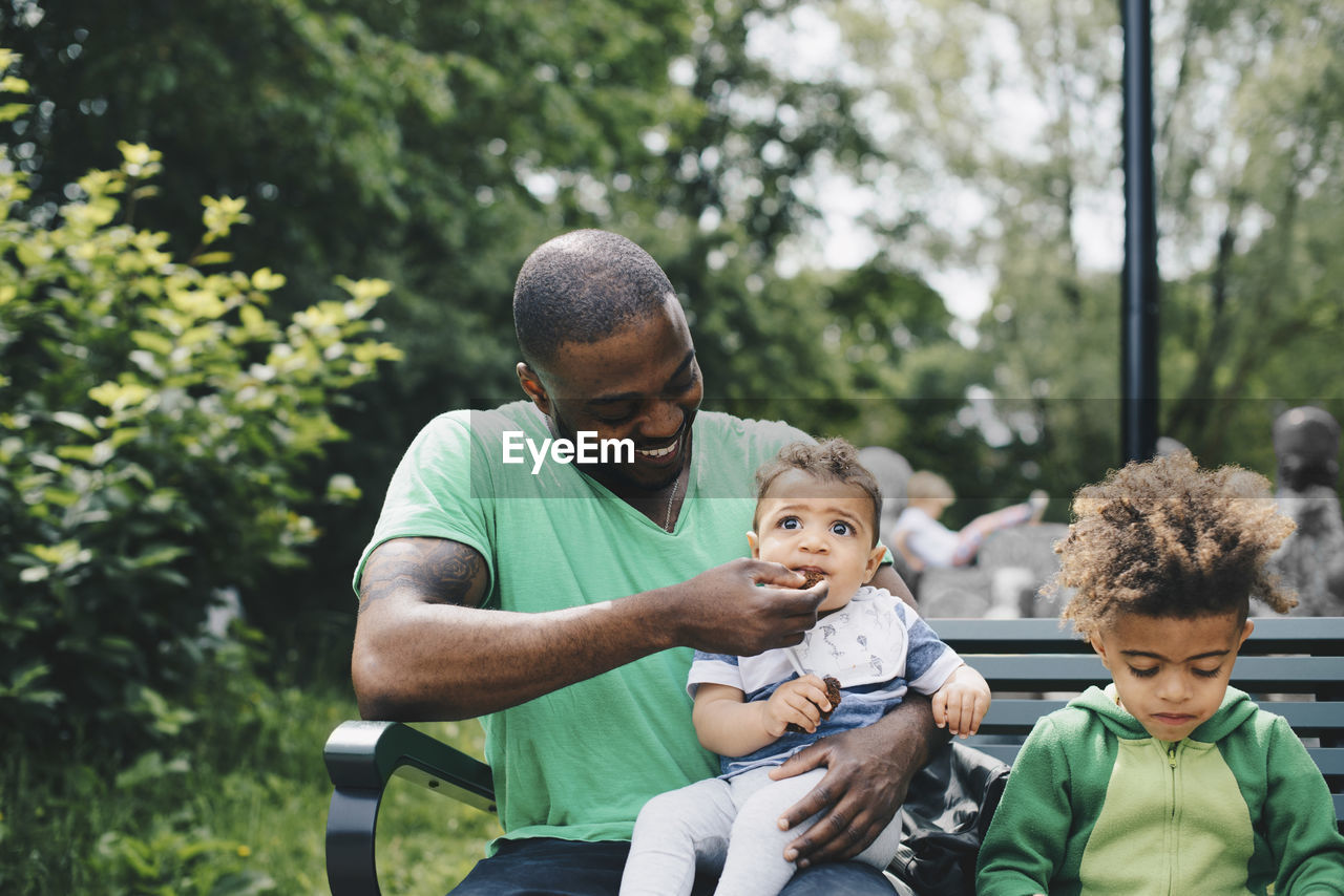 Smiling father feeding baby boy while sitting with son on bench at park