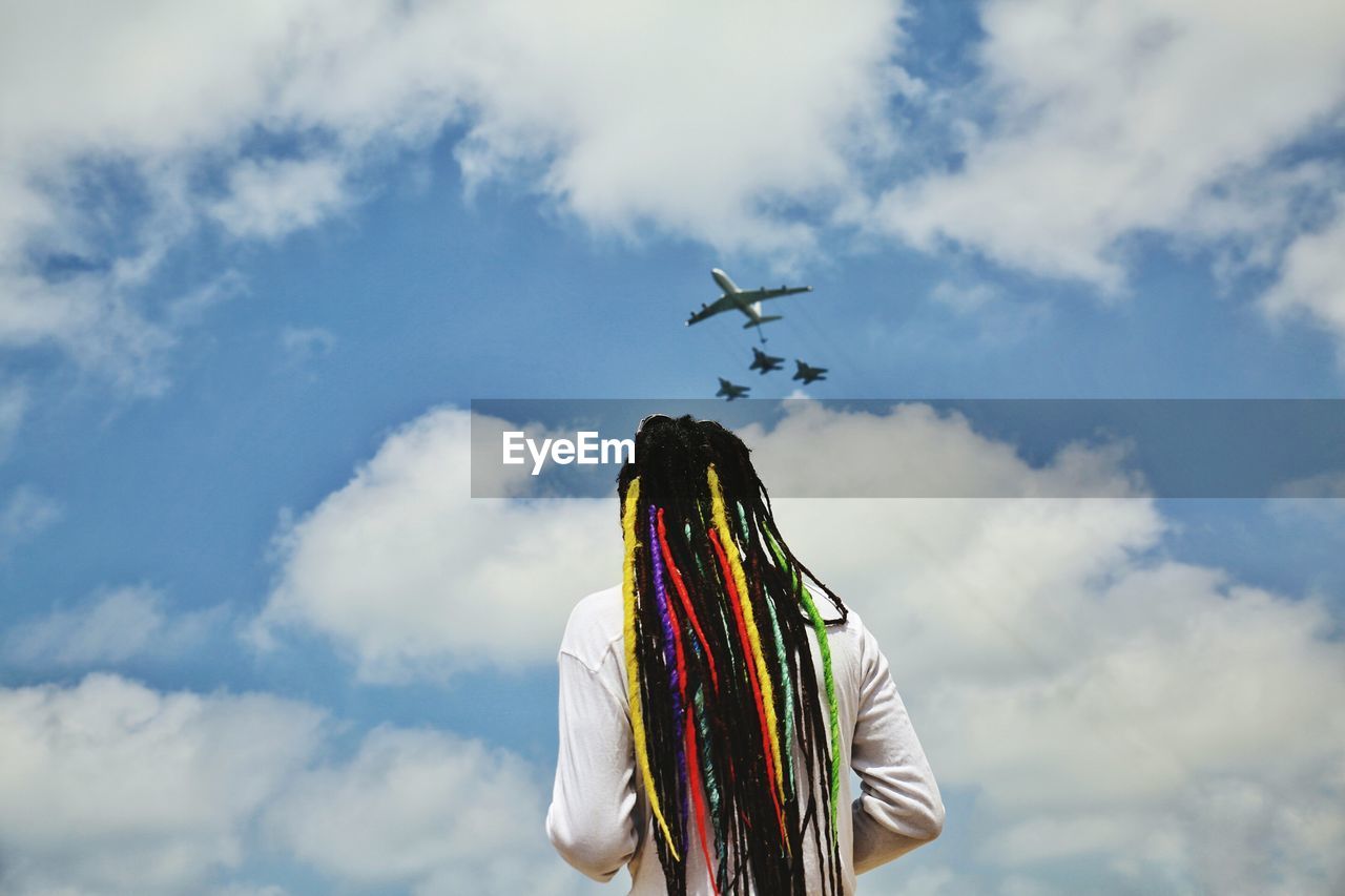 Rear view of man with colorful dreadlocks looking at airplanes flying against cloudy sky