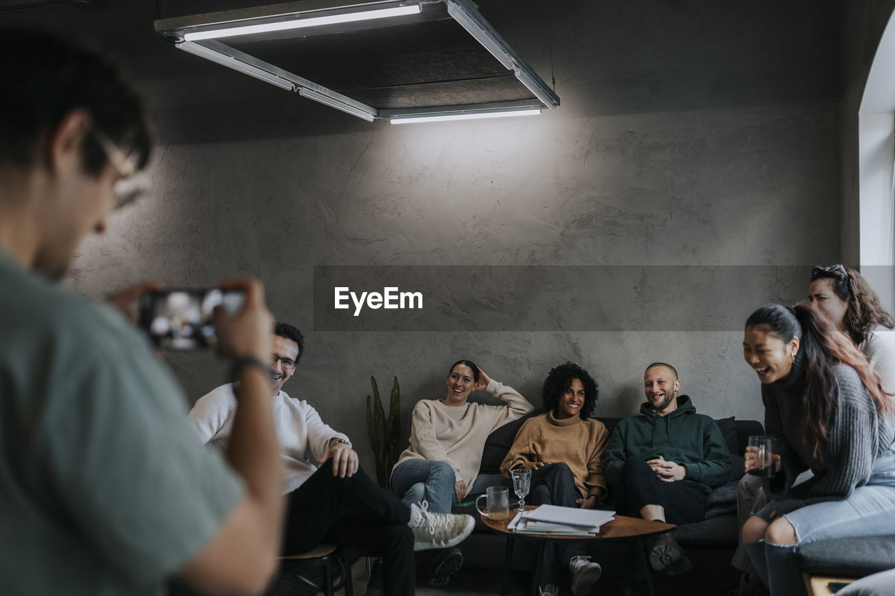 Male entrepreneur photographing happy colleagues through mobile phone at startup company