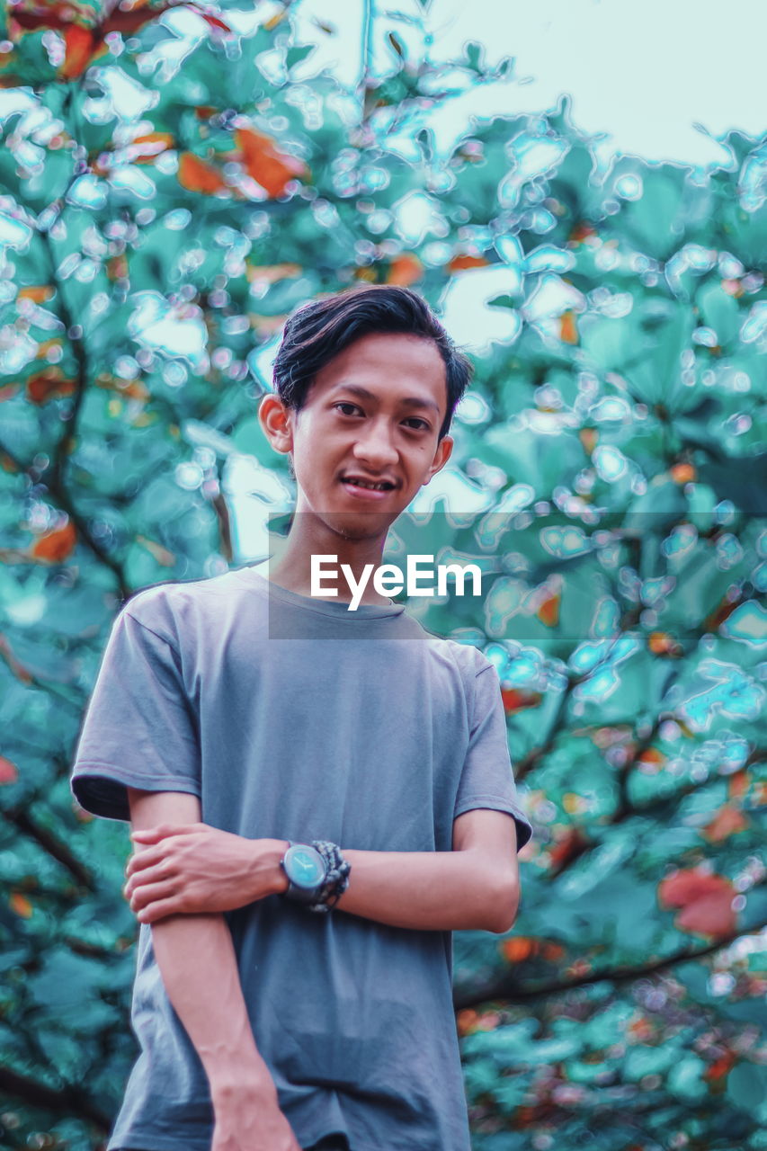 PORTRAIT OF SMILING YOUNG MAN STANDING AGAINST TREES