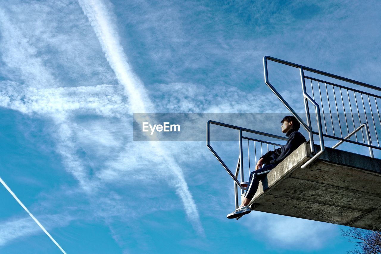 Low angle view of man sitting on built structure against blue sky