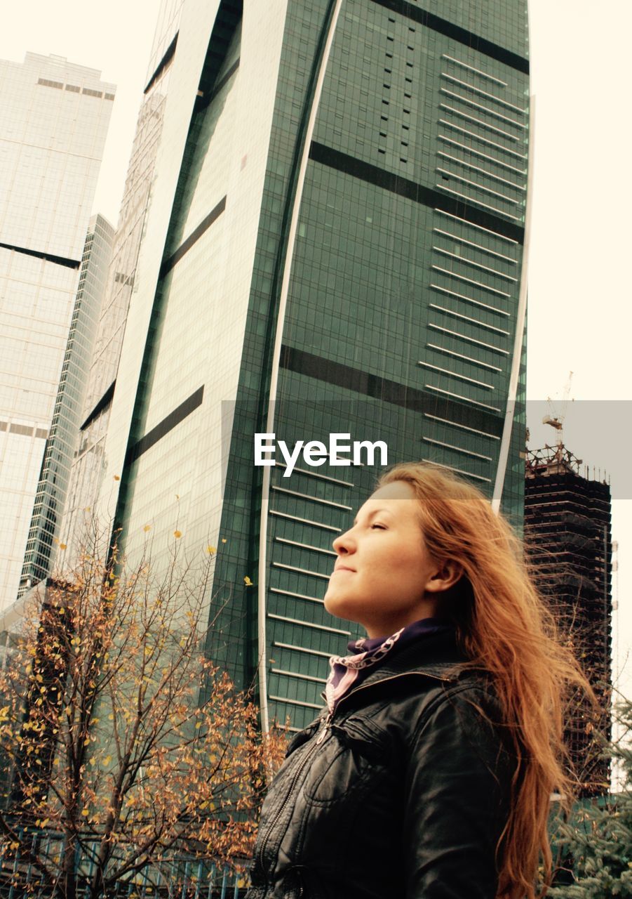 Low angle view of young woman standing with eyes closed against modern buildings