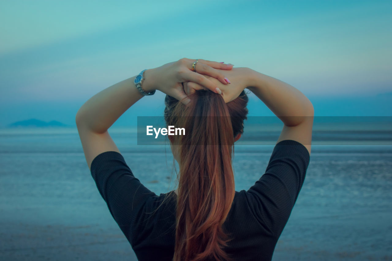 Rear view of woman with hands behind head standing at beach against sky