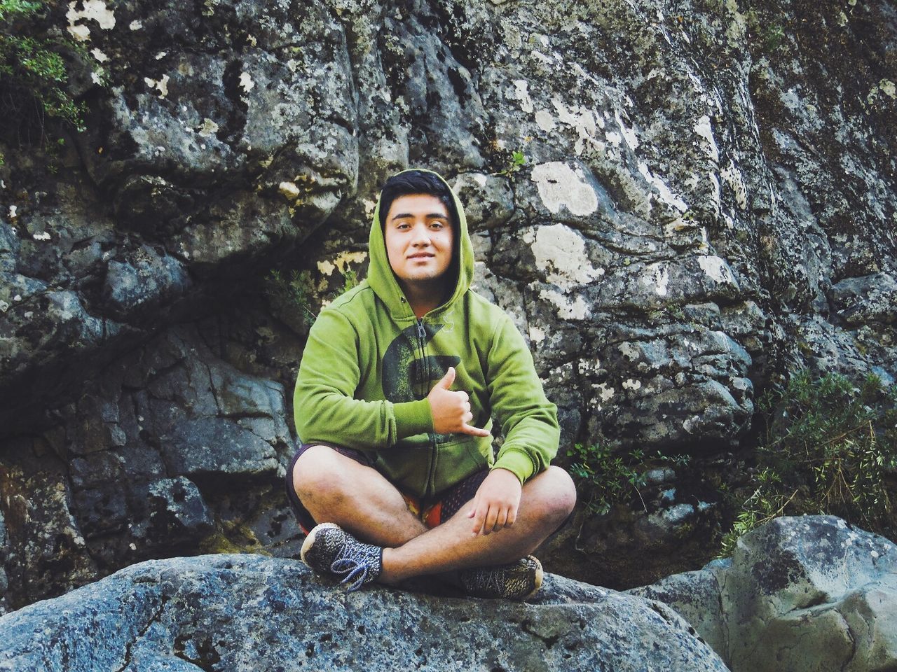 Full length portrait of man showing shaka sign while sitting on rock