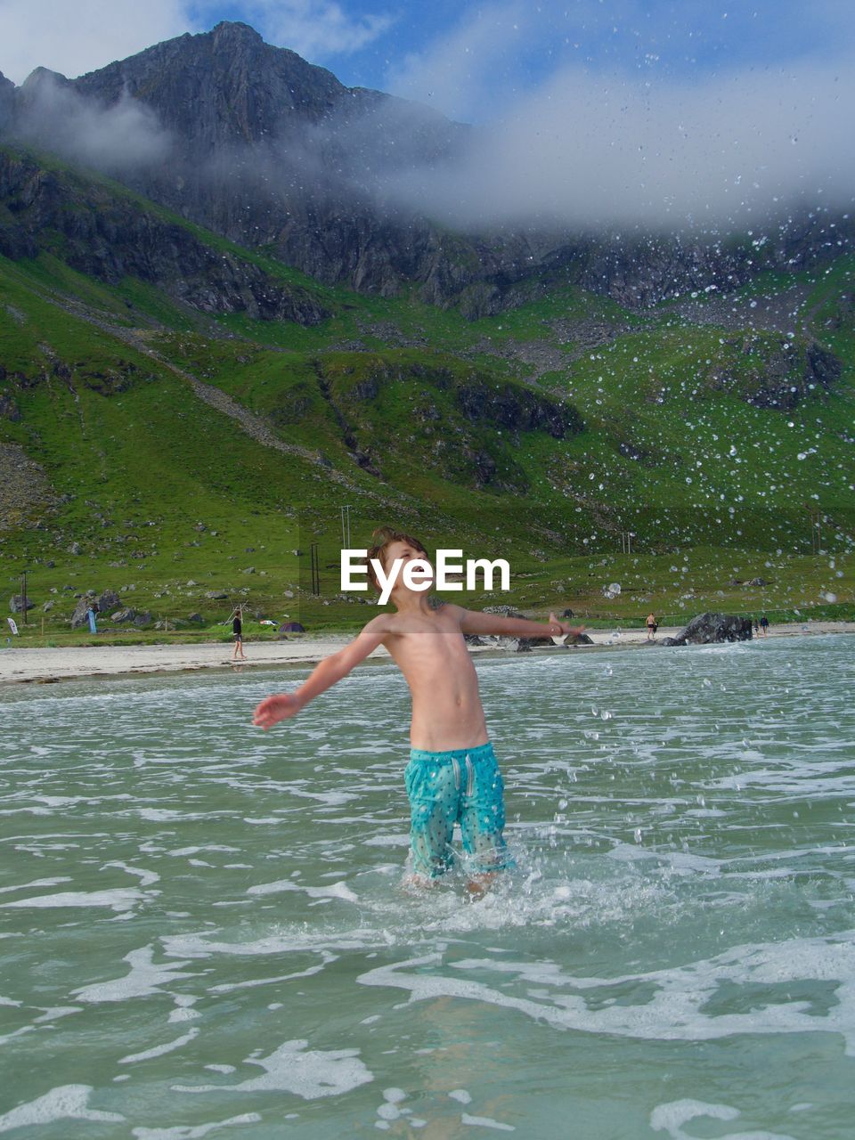 Shirtless boy with arms outstretched standing in sea against mountains