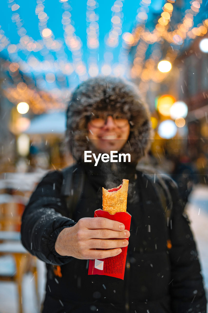 Smiling man eating warm pie on snowy city street in winter