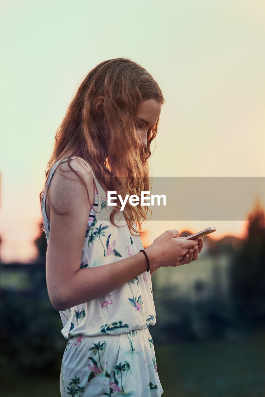 Side view of girl using mobile phone against sky during sunset