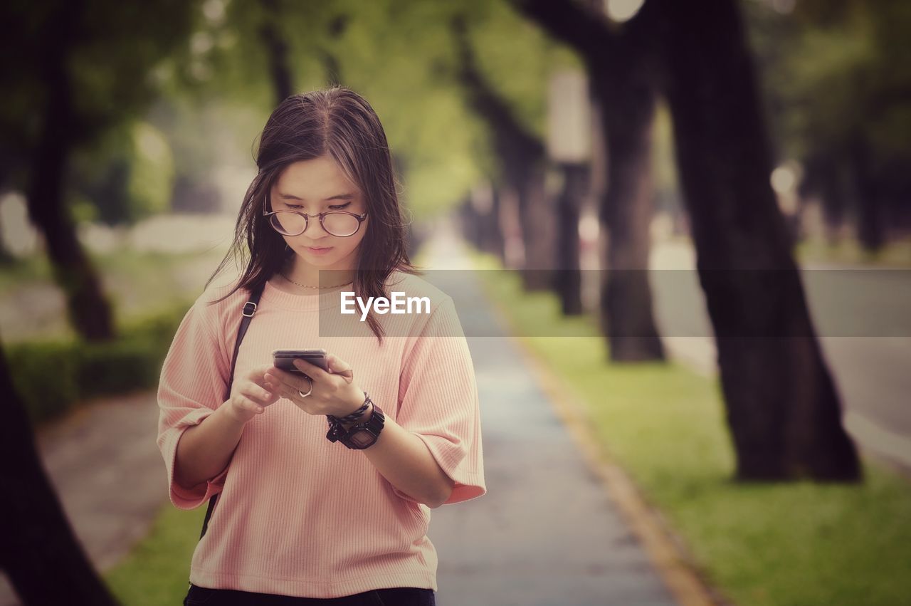 Young woman using smart phone while standing on footpath against trees in park