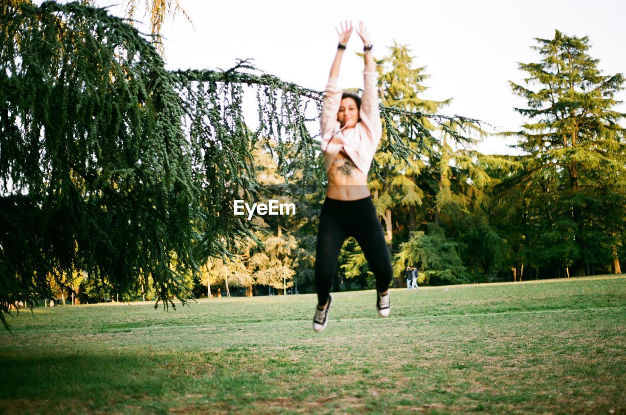 FULL LENGTH OF YOUNG WOMAN WITH ARMS RAISED IN PARK