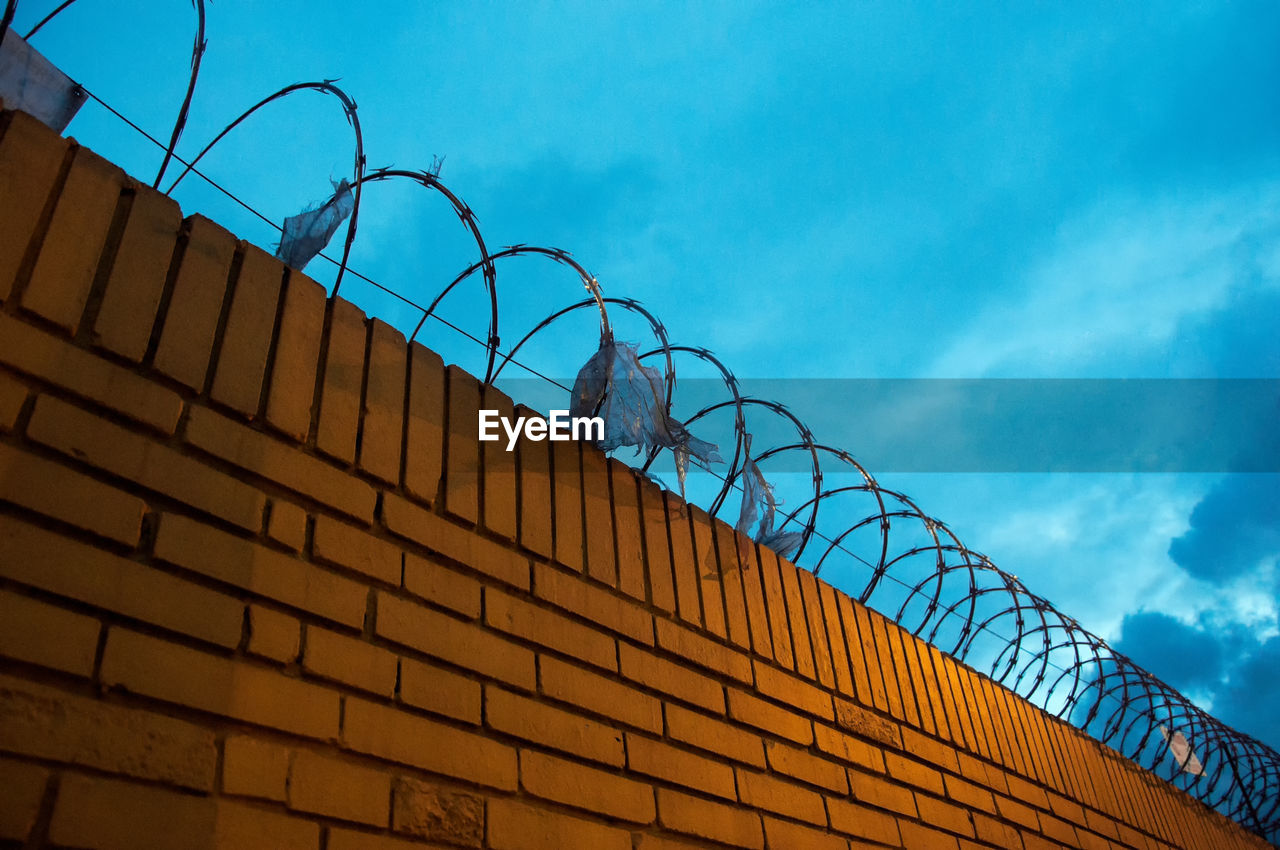 Low angle view of razor wire fence on brick wall against cloudy sky