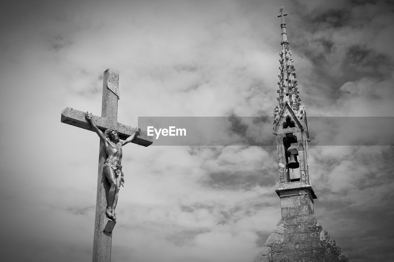 Low angle view of cross fix statue by bell tower against cloudy sky