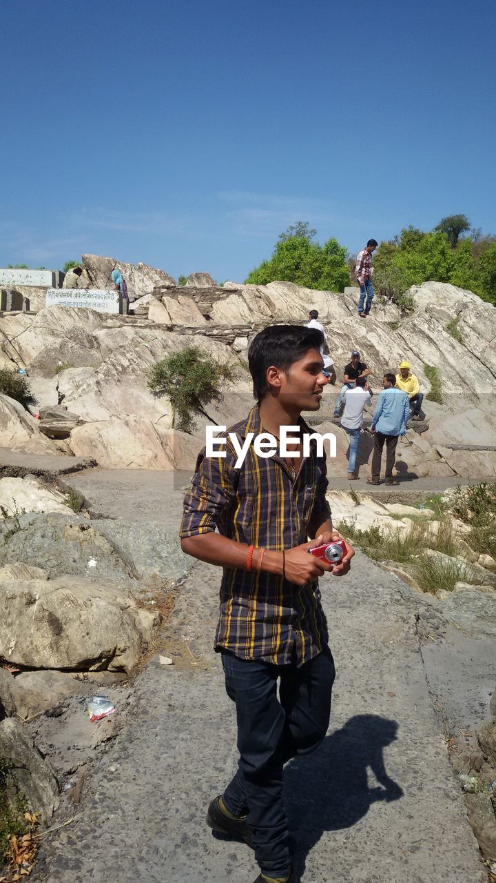 Man with camera looking away while standing on footpath against sky