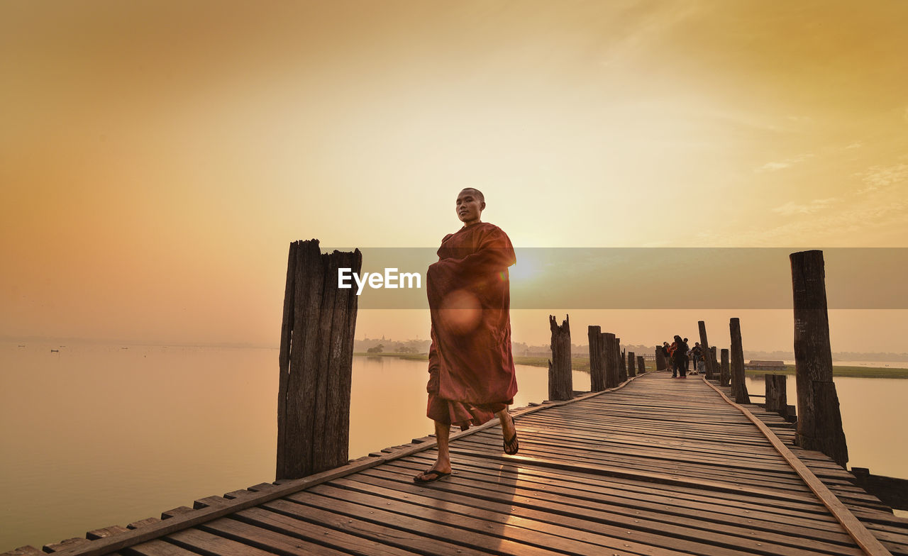 Full length of monk walking on pier over sea against sky during sunset