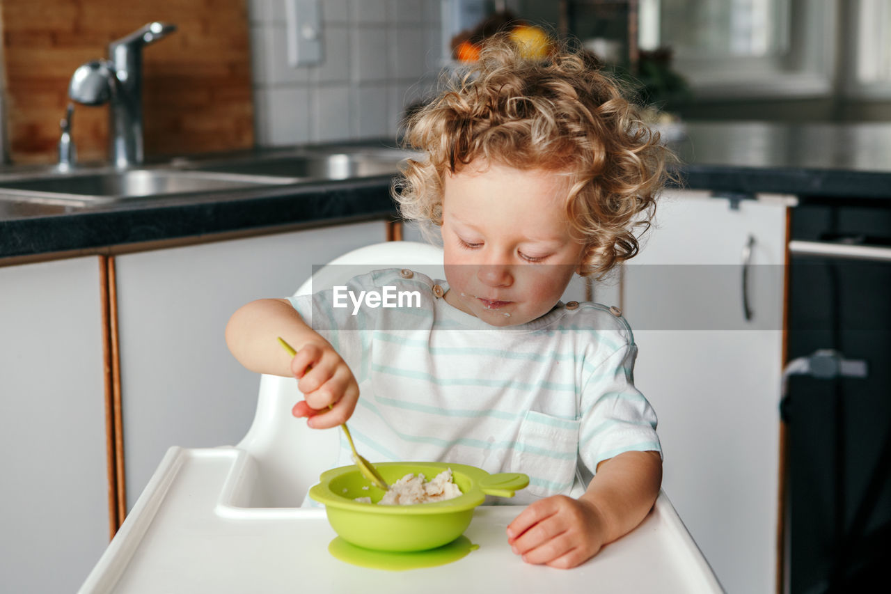 Caucasian curly kid boy sitting in high chair eating cereal puree with spoon. healthy eating 
