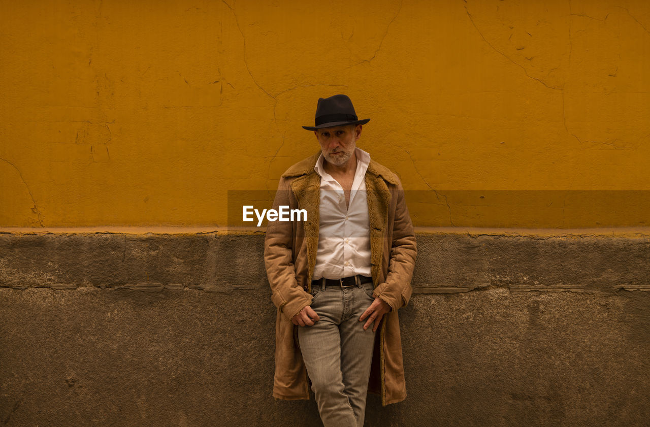 Portrait of adult man in hat and coat against yellow wall on street