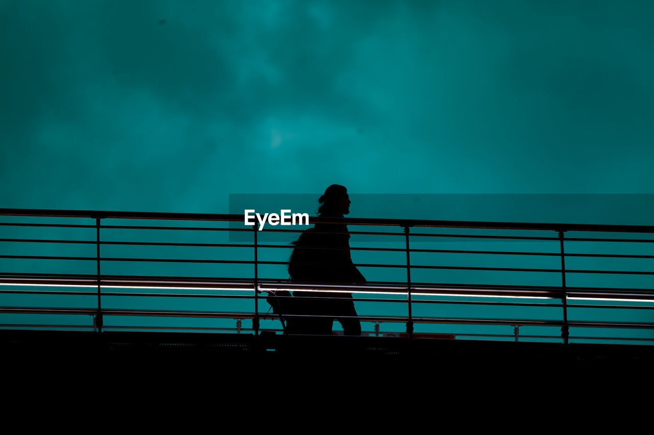 Low angle view of silhouette man walking by railing against sky