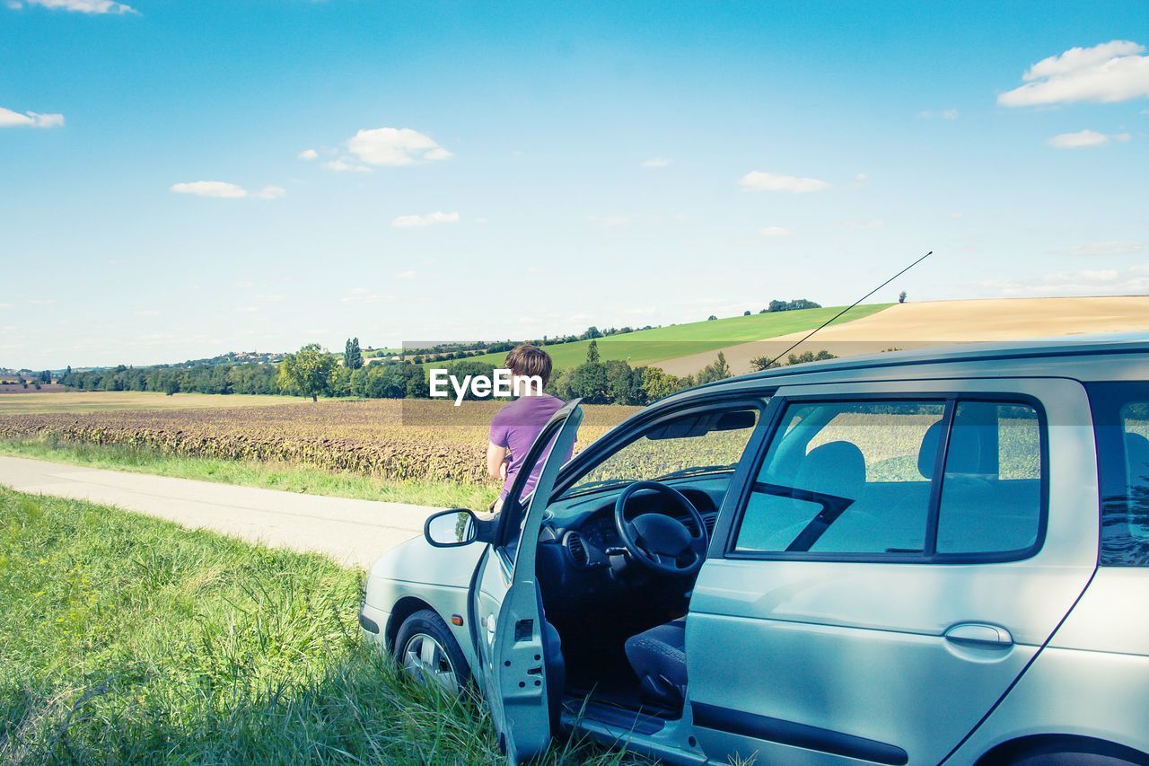 Man sitting on car hood against cloudy sky