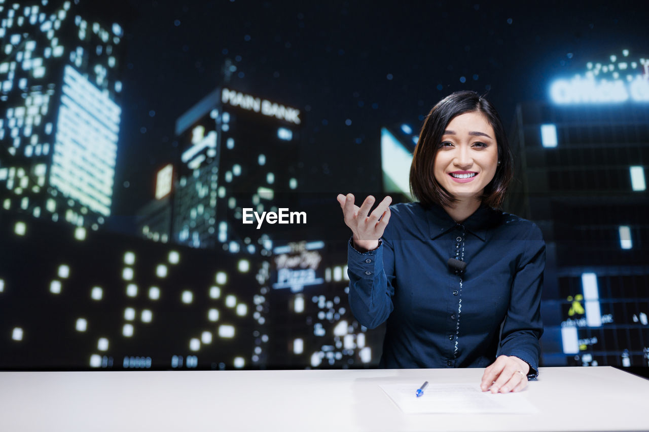 portrait of young woman using mobile phone while standing against illuminated city at night