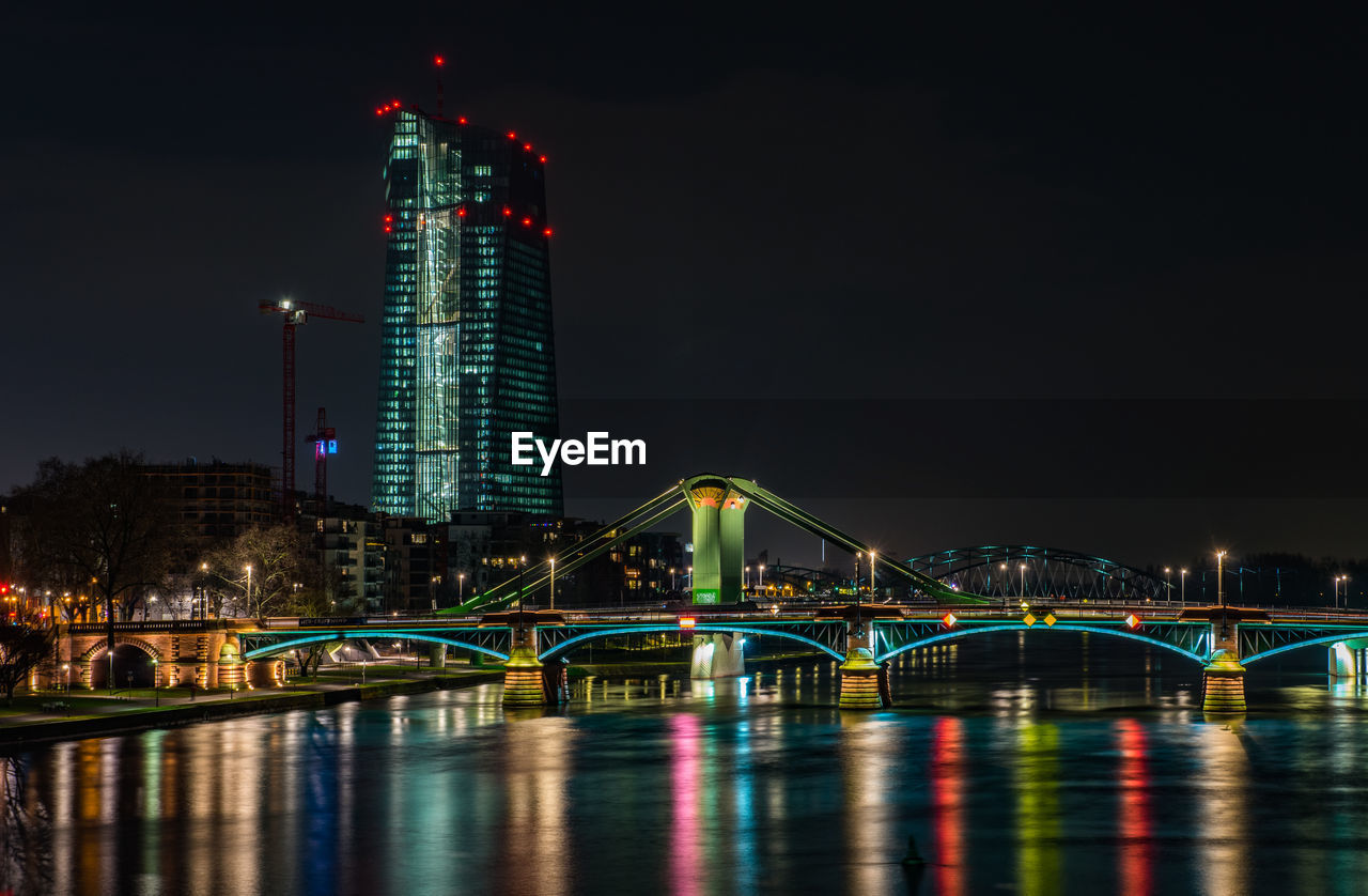 Illuminated bridge over river against buildings at night