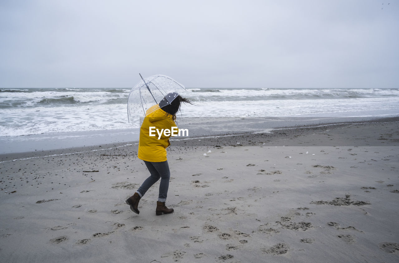 Woman in raincoat walking with umbrella at sea shore against sky