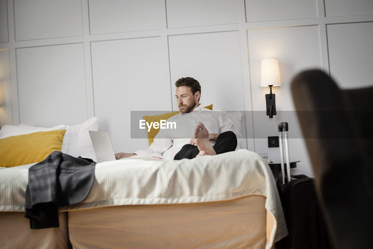 Businessman sitting on bed using laptop against wall in hotel room