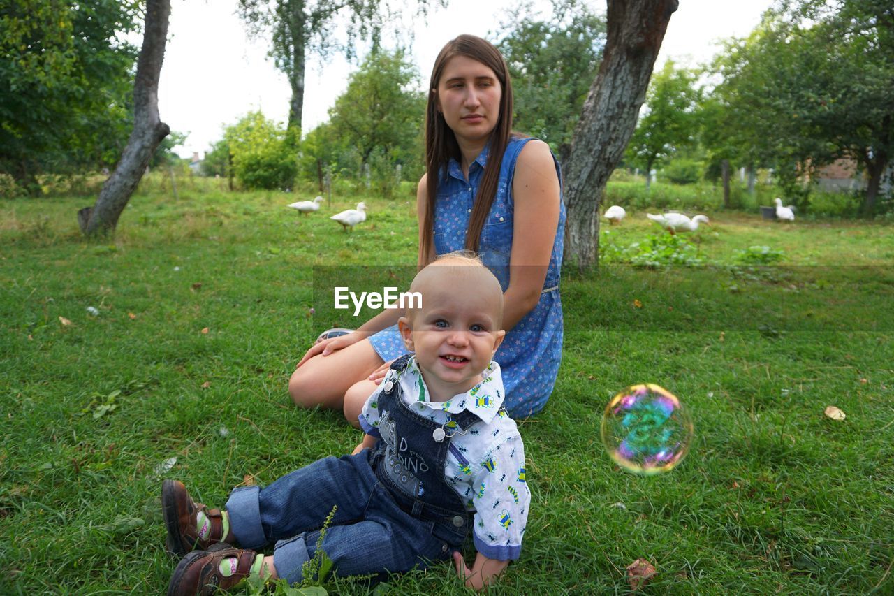 Portrait of boy with mother sitting by bubble on grassy field at park