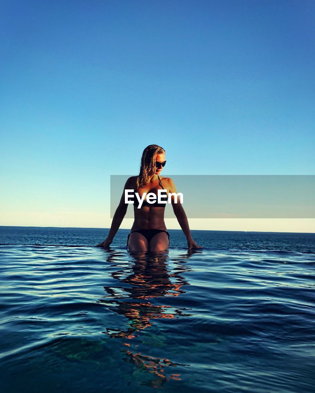 Sensuous woman in infinity pool by sea against clear blue sky